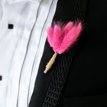 Closeup view of groom boutonniere with vibrant pink artificial feather design wrapped in jute placed on black suit for wedding.