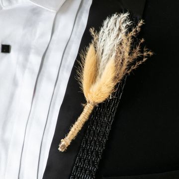 Closeup of boutonniere with beige and white dried grass stems wrapped in jute placed on black suit lapel.