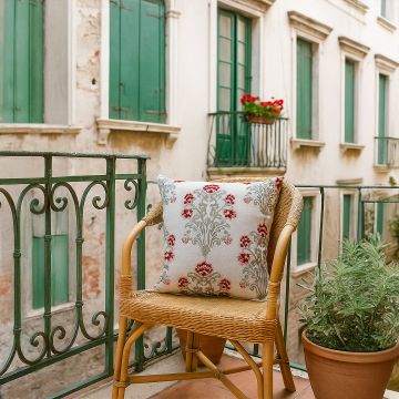 Cotton cushion with red and grey embroidered floral motif placed on a wicker chair in a balcony setting creating a warm cozy lifestyle decor accent for homes.