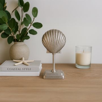 A decorative seashell figurine on a stand displayed on a wooden table with a candle green leaves and coastal themed books against a white background.