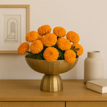 Table decor scene showing a golden pedestal bowl filled with bright orange marigold flowers placed on a wooden console with a beige vase and framed artwork in the background.