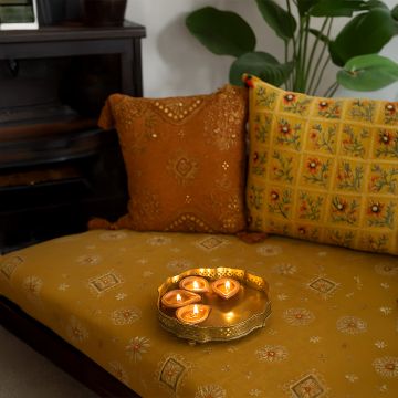 Warm festive scene with a golden diya tray holding glowing tealights placed on an embroidered mustard seat with decorative cushions in a cozy home setting.