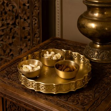 A decorative gold serving tray with matching bowls filled with assorted snacks displayed on an intricately carved wooden table creating a warm traditional setting.