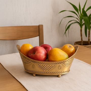 A decorative golden fruit basket filled with fresh apples and mangoes placed on a wooden dining table with a neutral table runner creating a warm and homely lifestyle setting.