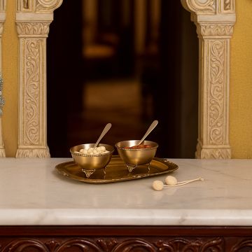 A brass serving set with two decorative bowls and spoons placed on an ornate golden tray displayed on a carved marble console framed by intricate traditional pillars creating a warm heritage lifestyle scene.