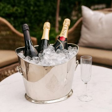 Lifestyle shot of a polished silver ice bucket filled with ice and three champagne bottles on an outdoor table creating an elegant celebration vibe.