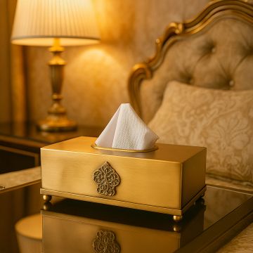 A gold tissue box with an ornate emblem placed on a reflective bedside table in a warm luxurious bedroom setting with soft ambient lighting.