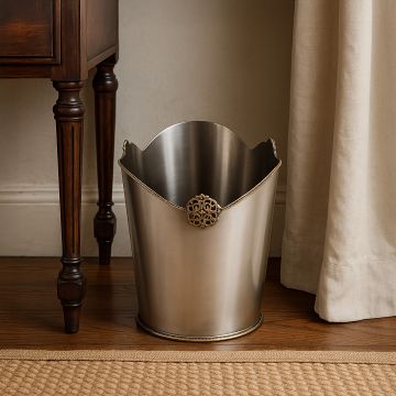 A luxury silver metal dustbin with ornate detailing placed beside a wooden table on a polished wooden floor adding elegance to a classic home interior.