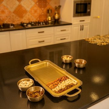 A rectangular gold serving tray displayed on a dark kitchen counter holding assorted nuts with matching gold bowls placed around it in a warm kitchen setting.