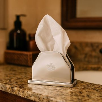 Silver plated napkin holder with engraved crest holding tissues on a granite bathroom counter next to soap bottles.