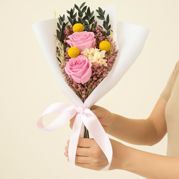 Lifestyle image of pink rose bouquet with white hydrangea yellow billy button and greenery wrapped in white paper with ribbon creating a preserved flower arrangement.
