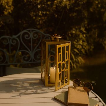 Golden metal lantern placed on a white outdoor table with sunlight reflections surrounded by books and sunglasses for a cozy garden setting.