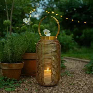 
Gold metal wire mesh lantern with circular handle illuminated with candle on outdoor garden patio stone path at dusk.