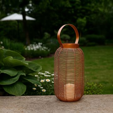 Copper metal wire mesh lantern with candle light glowing on stone patio edge beside green hosta plants and white daisies.