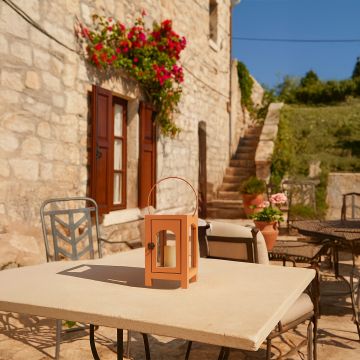 A warm outdoor lifestyle setting featuring a peach lantern placed on a stone table with blooming flowers and rustic stone walls creating a cozy natural ambiance in soft sunlight.