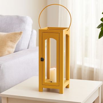 Bright yellow metal candle lantern with handle placed on a red brick garden path amidst green plants and flowers.