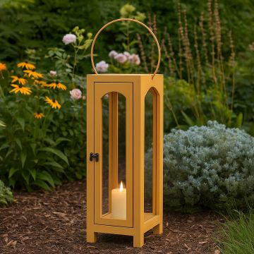 Natural wood floor lantern with a lit candle placed in a garden bed surrounded by plants creating a cozy warm outdoor ambiance.