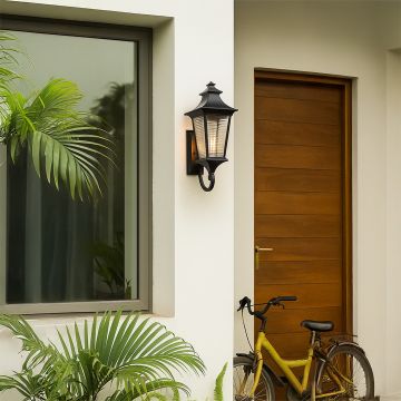 Classic black outdoor lantern wall light with ribbed glass illuminated on a white stucco wall beside a wooden door and window with plants.
