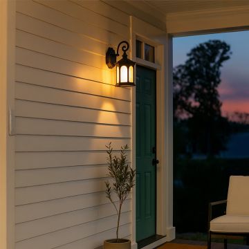 Classic black outdoor lantern wall light with ribbed glass illuminated on a white stucco wall beside a wooden door and window with plants.