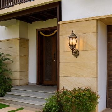 Antique gold outdoor wall lantern illuminated on beige stone wall near dark wood door with traditional marigold garland.