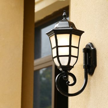 Black metal outdoor wall lantern light on with frosted glass panels mounted beside a window creating warm ambient lighting for porch patio and home entrance Decor.
