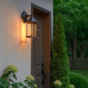 An outdoor wall lantern with the light on illuminating the entrance pathway and garden plants creating a warm welcoming glow beside a modern exterior door at dusk.