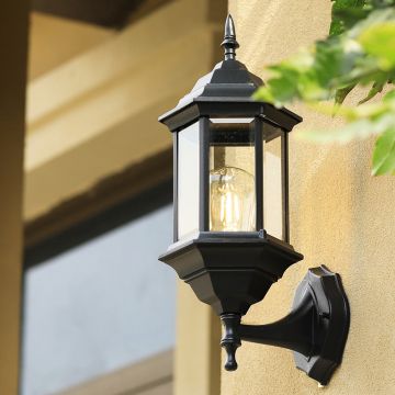 Modern black outdoor lantern mounted on a wall with raindrop glass casting a warm ambient glow in a simple lifestyle setting.