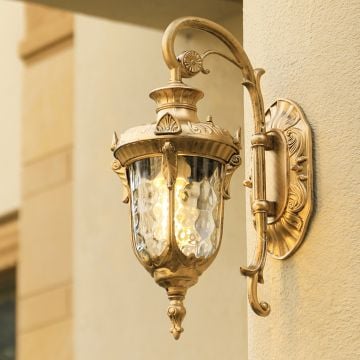 A gold antique outdoor wall lantern with the light on illuminating the textured wall and creating a warm elegant glow that enhances the classic architectural exterior.