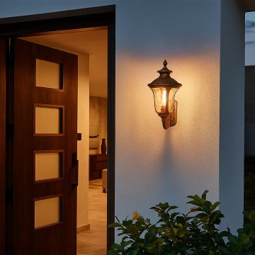 A gold vintage outdoor wall lantern with the light on illuminating the entrance wall and casting a warm welcoming glow that enhances the exterior ambiance near the doorway at dusk.