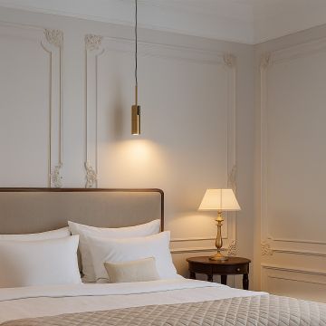 Modern gold cylindrical pendant light glowing above bed elegant ceiling lamp for luxury bedroom lighting and stylish interiors.
