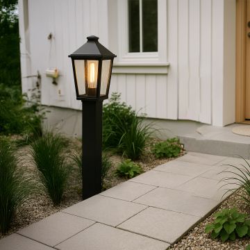A modern outdoor pathway scene featuring a tall black post light turned on casting a warm glow beside a stone walkway near a white house entrance at dusk.