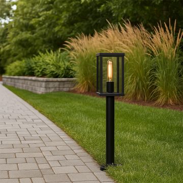 A black garden bollard light turned on beside a paved path casting a warm glow over green lawn and plants at dusk creating an inviting outdoor scene.
