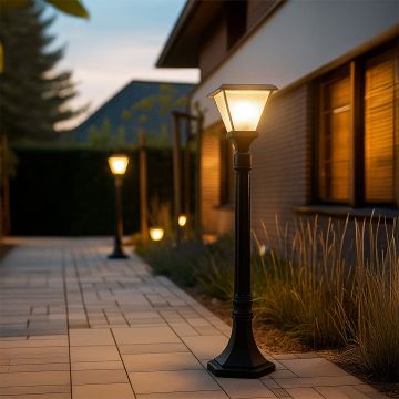 A black outdoor pathway lamp post with the light on illuminating a modern garden walkway at dusk creating a warm inviting glow along the landscaped path and house exterior.