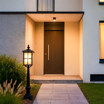 A classic outdoor pillar lamp with the light on illuminating the entry walkway and casting a warm glow across the garden plants creating an inviting ambiance near the front door.