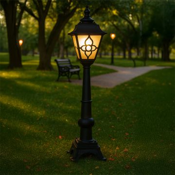A classic outdoor pathway lamp with the light on glowing warmly in a green park setting and illuminating the grass and walking path to create a calm inviting evening atmosphere.