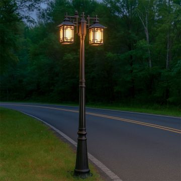 Twin head black outdoor post light with glowing frosted lanterns lighting a roadside path at dusk creating warm ambient illumination in a natural outdoor setting.