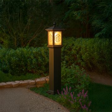 Brown outdoor pillar lamp with glowing decorative Chinese pattern glass lighting a garden pathway at night creating a warm inviting ambience among lush greenery.