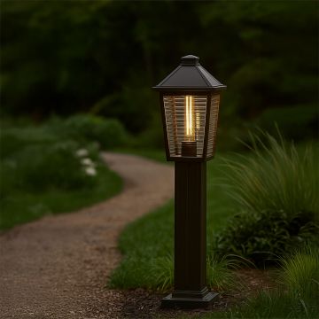 A black outdoor pillar lamp with ribbed glass glowing warmly along a garden pathway at night creating a soft inviting ambience surrounded by lush greenery.