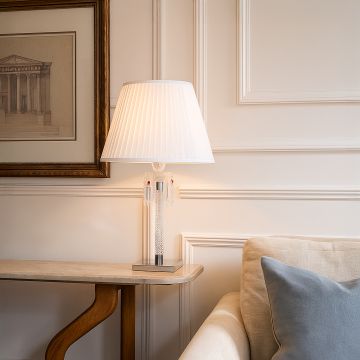 Glass lamp with white pleated shade glowing warmly placed on a side table beside sofa and framed wall art in living room.