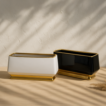 A pair of modern rectangular ceramic planters in white and black with gold bases placed on a wooden surface in soft natural light creating a warm decorative look.
