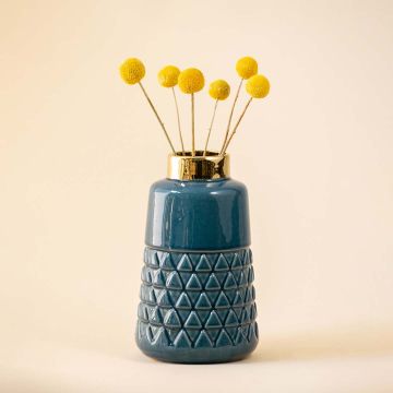 A blue ceramic vase with a gold rim and geometric pattern is filled with yellow craspedia flowers and placed on a light colored surface.