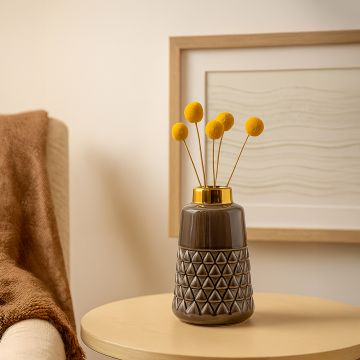 A modern ceramic vase with a textured geometric base and golden top displayed on a wooden side table beside cozy home decor creating a warm lifestyle setting.