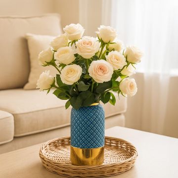 A blue textured ceramic vase with a gold base holding fresh white roses placed on a woven tray in a bright cozy living room setting.