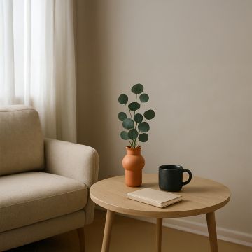 Modern living room setup featuring a ceramic orange vase with green eucalyptus leaves on a light wood round table beside a beige sofa.