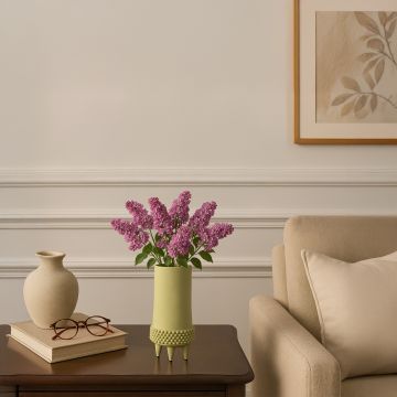 Light green ceramic vase with textured base holding purple flowers placed on wooden side table beside beige sofa.