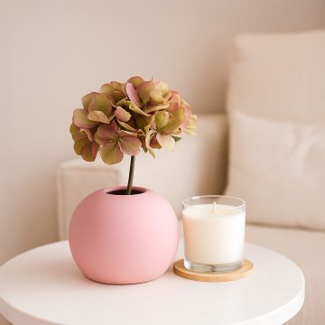 Pink round vase with a single hydrangea stem placed on a white table beside a glass candle  styled in a soft and cozy living room setting.