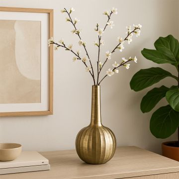 Elegant brass vase with a tall narrow neck and rounded base holding delicate white blossom stems placed on a light wooden table beside soft decor accents.