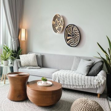 A handwoven natural and black rattan wall decor plate styled above a grey sofa with wooden tables indoor plants and soft neutral accents creating modern warm home vibes.