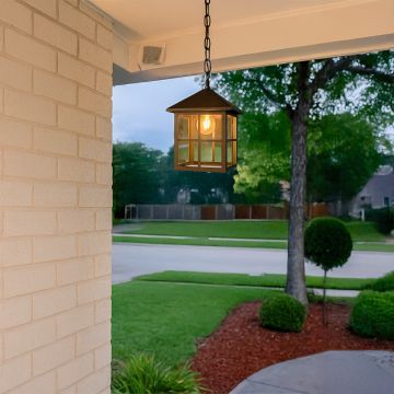 Black metal round cage pendant light hanging from a modern pergola ceiling with warm light on against a blue evening sky.