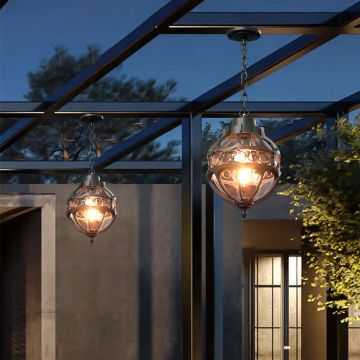 Black metal round cage pendant light hanging from a modern pergola ceiling with warm light on against a blue evening sky.